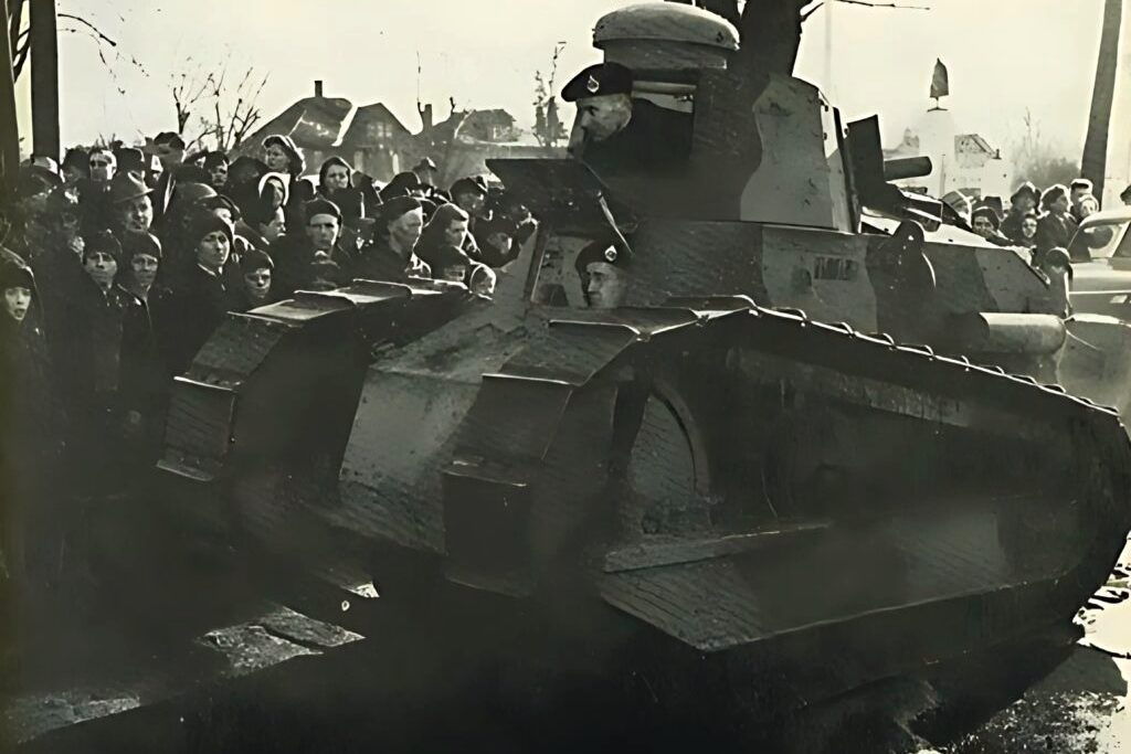 A Renault M1917 light tank driving down an Oshawa street in 1940, its commander in the open turret hatch and a crowd of onlookers pressed along the kerb behind it.